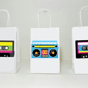 May include: Three white paper gift bags with handles. Each bag has a different retro-themed design: a cassette tape with green, yellow, and pink stripes, a blue boombox with black and yellow accents, and a cassette tape with pink, yellow, and blue stripes.