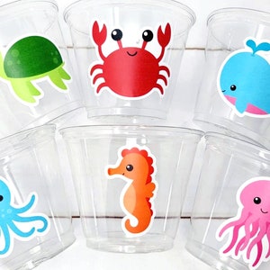 May include: Six clear plastic cups with colorful sea creature stickers. The cups feature a green turtle, a red crab, a blue whale, a blue octopus, an orange seahorse, and a pink jellyfish.