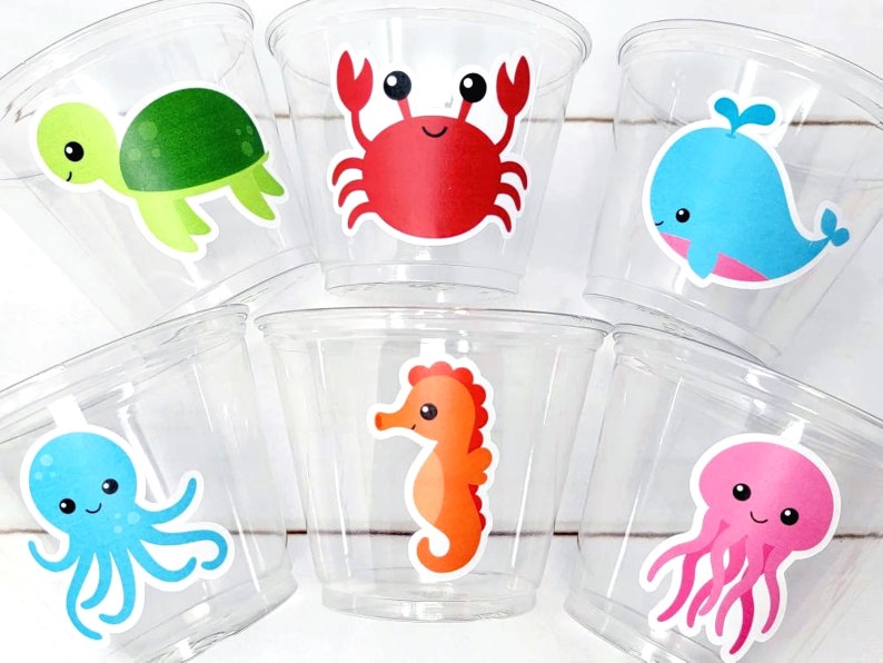 May include: Six clear plastic cups with colorful sea creature stickers. The cups feature a green turtle, a red crab, a blue whale, a blue octopus, an orange seahorse, and a pink jellyfish.