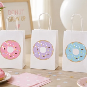 May include: Three white paper gift bags with twisted paper handles, each featuring a large donut graphic. The donuts are pink, light purple, and light blue, all decorated with colorful sprinkles. The bags stand upright in a row on a flat surface.