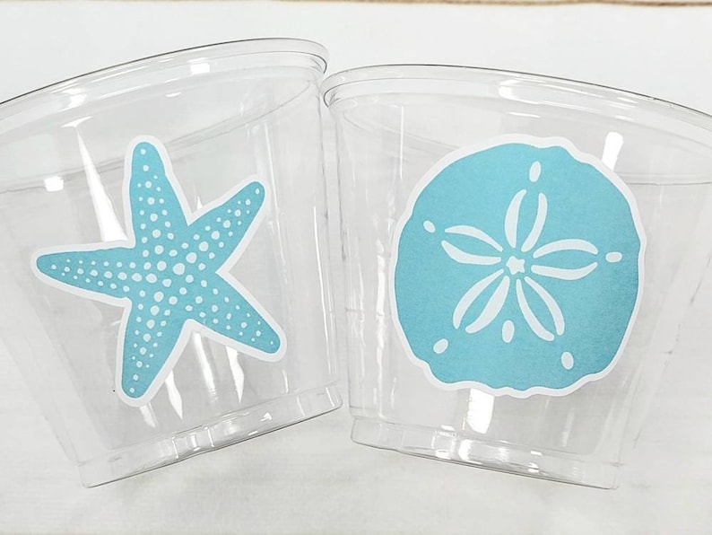 May include: Two clear plastic cups with blue and white starfish and sand dollar designs. The cups are perfect for a beach-themed party.