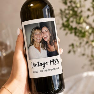 May include: A dark glass wine bottle featuring a custom rectangular label with a photograph of two smiling individuals. The label includes the black printed text Vintage 1986 and AGED TO PERFECTION in a combination of script and sans-serif fonts.