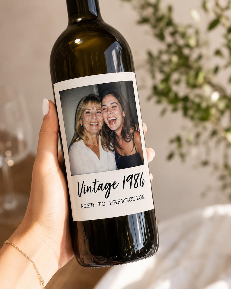 May include: A dark glass wine bottle featuring a custom rectangular label with a photograph of two smiling individuals. The label includes the black printed text Vintage 1986 and AGED TO PERFECTION in a combination of script and sans-serif fonts.