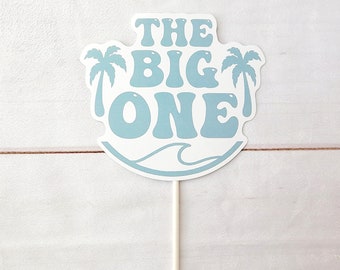 May include: Blue and white cake topper with the text "THE BIG ONE" and palm trees on either side. The topper is on a white stick.