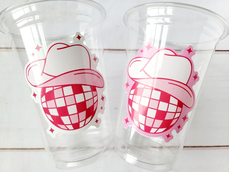 May include: Two clear plastic cups, each decorated with a pink and white graphic. The graphic features a cowboy hat above a red and white checkered disco ball, with pink starbursts around it.