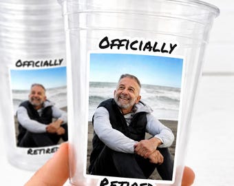 Könnte beinhalten: Transparente Plastikbecher mit einem Aufkleber mit der Aufschrift "Officially Retired" und einem Foto eines lächelnden Mannes. Die Becher werden in einer Hand gehalten, mit weiteren Bechern im Hintergrund. Das Foto zeigt den Mann am Strand sitzend.