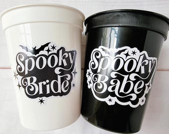 May include: Two plastic cups, one white and one black, with Halloween-themed designs. The white cup says "Spooky Bride" in a black, stylized font with bat and star accents. The black cup says "Spooky Babe" in a similar style.