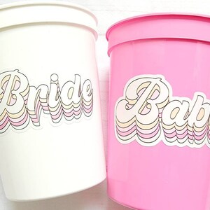 Puede incluir: Dos vasos de pl&aacute;stico con pegatinas de estilo retro. Un vaso es blanco con la palabra "Bride" en rosa, amarillo y blanco. El otro vaso es rosa con la palabra "Babe" en rosa, amarillo y blanco.