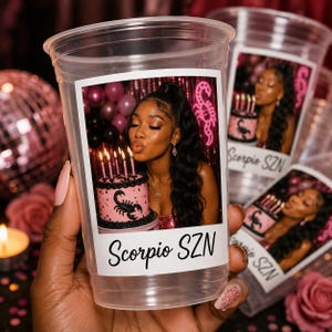 May include: A clear plastic disposable cup featuring a rectangular photo print of a person blowing out candles on a pink cake with a black scorpion graphic. The text Scorpio SZN is printed in black script below the photo on the cup surface.