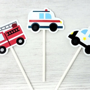 May include: Set of three vehicle-themed cupcake toppers. Includes a red fire truck, a white ambulance with red stripes, and a black and white police car with a yellow star. Each topper is attached to a white stick.