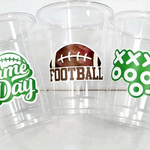 May include: Three clear plastic cups with green and white designs. The first cup has a green and white football design with the text "FOOTBALL". The second cup has a green and white design with the text "game Day". The third cup has a green and white design with the text "XXX" and three circles.