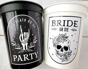 May include: Two black and white plastic cups with a skull and floral design. One cup says "Bride or Die" and the other says "Til Death Do Us Party".
