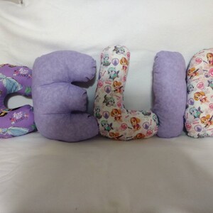 May include: Pillows spelling out the name "CELIA". The letters are in various patterns, including a purple fabric with princess characters and a white fabric with Paw Patrol characters. The letter "E" is a solid lavender color.