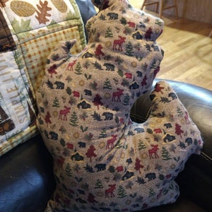 May include: A decorative pillow with a unique shape, featuring a pattern of bears, moose, and trees in shades of red, green, and black on a gray background. The pillow is placed on a black leather couch.