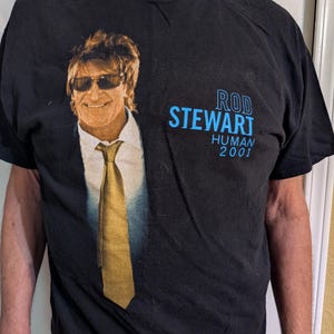 May include: Black t-shirt featuring a graphic of Rod Stewart wearing sunglasses and a suit with a gold tie. The text "ROD STEWART HUMAN 2001" is printed in blue on the right side of the shirt.