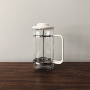 Op de afbeelding: Een glazen cafetière met een witte handgreep, deksel en rand. De cafetière heeft een roestvrijstalen filter en een donkerbruin houten oppervlak.