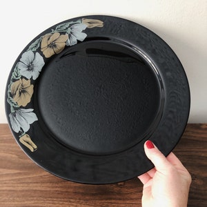 Puede incluir: Un plato de cena negro con un diseño floral alrededor del borde. Las flores son en tonos de amarillo, verde y blanco.