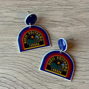 May include: A pair of novelty earrings featuring a design with a rainbow, mountains, and the text "USCSS NOSTROMO 180286". The earrings have a blue stud and a white border.