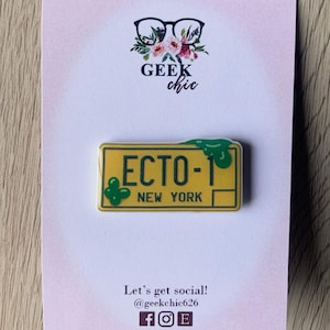 May include: A yellow and green enamel pin featuring a license plate with the text "ECTO-1 NEW YORK". The pin is on a white background with the text "GEEK chic" and social media handles.