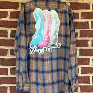 May include: A long-sleeved, button-down flannel shirt with a brown, blue, and white plaid pattern. The back of the shirt features a graphic of three women in different colors with the text "Britney Spears" in white script.