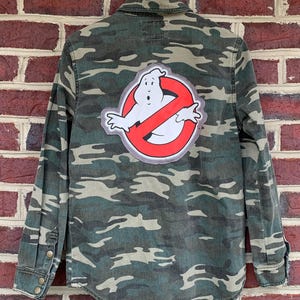May include: A green and brown camouflage button-up shirt with a Ghostbusters "No Ghost" logo patch on the back.