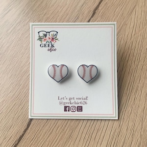 May include: A pair of heart-shaped stud earrings with a baseball design. The earrings are white with red stitching and a blue outline. The earrings are on a white card with the text "Geek chic" and "Let's get social! @geekchic626 f o e".