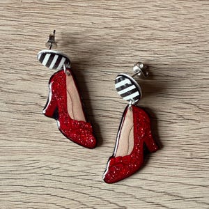 May include: A pair of dangle earrings featuring red glitter high heels with a black and white striped accent. The earrings have a post back and are set against a light brown wooden surface.