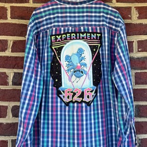 May include: Blue, pink, and green plaid button-down shirt with a patch featuring a cartoon character inside a glass dome. The patch reads "EXPERIMENT" and "626". The shirt is hanging on a black hanger against a brick wall.