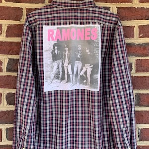 May include: A blue and red plaid long-sleeved button-up shirt with a Ramones band patch on the back. The patch features a black and white photo of the band members and the band name in pink.