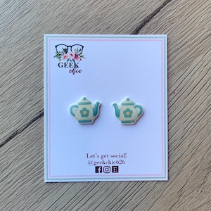 May include: A pair of teapot-shaped stud earrings on a white card with a pink and green border. The earrings are cream-colored with teal accents and a teal flower design. The card has the text "GEEK chic" and social media information.
