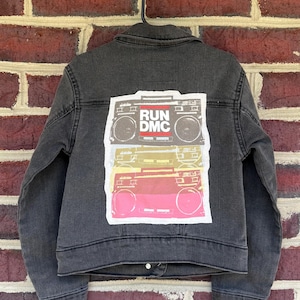 May include: A dark gray denim jacket with a graphic of three stacked boomboxes in red, gray, yellow, and pink. The top boombox has the text "RUN DMC" in red and white. The jacket is hanging on a black hanger.