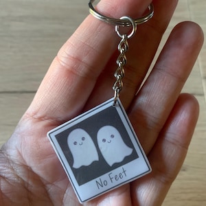 May include: A white keychain with two cartoon ghosts with smiling faces and the text "No Feet".