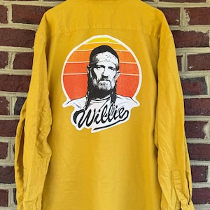 May include: Mustard yellow long-sleeve shirt with a button-down front. The back features a circular graphic of a man with a bandana and the name "Willie" in script. The graphic has orange and yellow sunset colors.