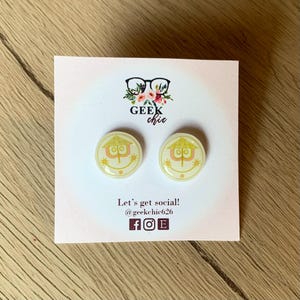 May include: A pair of yellow and pink stud earrings featuring a stylized smiley face with a house-like shape. The earrings are on a white card with the text "GEEK chic" and "Let's get social! @geekchic626 f o E".