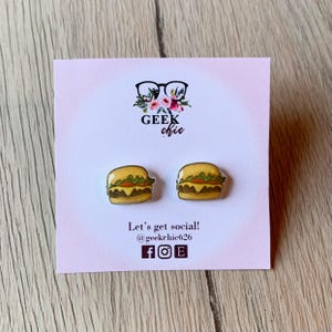 May include: A pair of small, yellow and brown hamburger earrings. The earrings are on a white card with the text "GEEK chic" and "Let's get social! @geekchic626" printed on it.