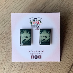 May include: A pair of earrings featuring a close-up image of a woman's face from the Scream movie poster. The earrings are rectangular and have a white background with the word "SCREAM" printed in black.