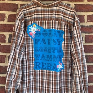 May include: Brown and white plaid button-down shirt with a blue patch on the back. The patch features the names Dolly, Patsy, Loretta, Tammy, and Reba in a stylized font, with floral accents.
