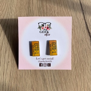 May include: A pair of yellow and black earrings featuring the text "ECTO-1" and "NEW YORK". The earrings are on a pink card with the text "GEEK chic" and "Let's get social! @geekchic626 fOF".