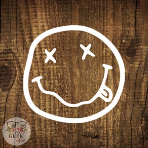 May include: White smiley face with crossed out eyes and a tongue sticking out, on a brown wooden background.