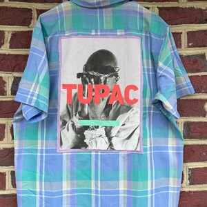 May include: A short-sleeved button-down shirt with a blue, green, and white plaid pattern. The back features a graphic of Tupac with the word "TUPAC" in red. The shirt is hanging on a black hanger.