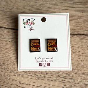 May include: A pair of black and gold Dungeon Crawler book-shaped earrings on a white card with a pink border. The card has the text "GEEK chic" and "Let's get social! @geekchic626".