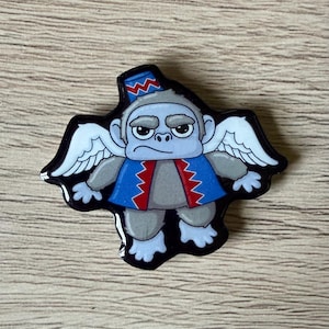 May include: A whimsical pin featuring a cartoon monkey with gray fur, wearing a blue and red jacket and a matching hat. The monkey has white wings and a stern expression. The pin is outlined in black.