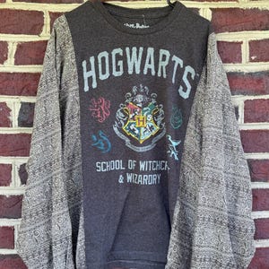 May include: Dark gray long-sleeve shirt with "HOGWARTS" printed on the front, along with the school crest and the words "SCHOOL OF WITCHCRAFT & WIZARDRY." The sleeves are patterned with a gray and white design. The shirt is hanging on a black hanger.