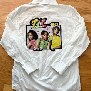 May include: White button-down shirt featuring a graphic of the music group TLC. The graphic includes the group's logo, the year 1992, and images of the three members. The shirt has long sleeves and a classic collar.