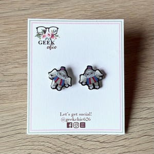 May include: A pair of stud earrings featuring a cartoon character with wings, wearing a red and blue outfit. The earrings are displayed on a white card with the text "GEEK chic" and social media handles.