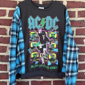 May include: Black sweatshirt with AC/DC graphic and "BLOW UP YOUR VIDEO" text. The sleeves are blue and black plaid. The graphic features a figure playing a guitar with neon green and pink accents.