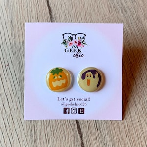 May include: A pair of round, beige earrings with Halloween-themed designs. One earring features a jack-o'-lantern, and the other has a ghost. The earrings are displayed on a white card with the text "Geek Chic" and social media information.