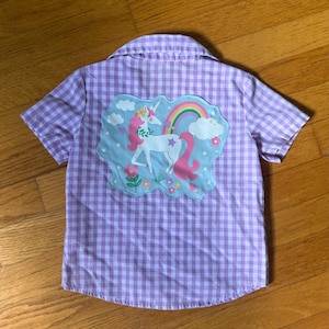 May include: A short-sleeved, button-down shirt with a lilac and white gingham pattern. The back features an appliqued design of a unicorn with a rainbow, clouds, and flowers. The unicorn has a pink mane and tail, and a gold horn.