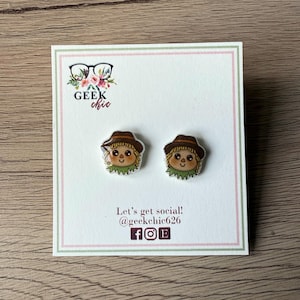 May include: A pair of stud earrings featuring a cartoon scarecrow design. The earrings are on a white card with the text "Geek Chic" and "Let's get social! @geekchic26". The scarecrows have brown hats, tan faces, and green collars.