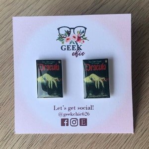 May include: A pair of Dracula book cover earrings. The earrings are made of resin and feature the classic cover art of the novel Dracula by Bram Stoker. The earrings are displayed on a white card with the text "Geek chic" and social media handles.
